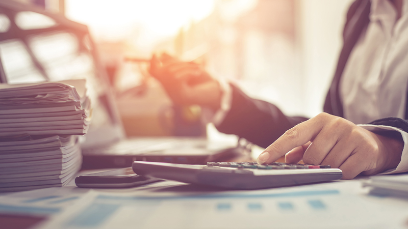Sobre um tampo de mesa, uma máquina calculadora simples é operada pela mão esquerda de uma pessoa vestindo fato, da qual só vemos parte do tronco, braços e mãos. Em segundo plano, a mão direita desta pessoa segura uma caneta. Em redor da máquina são visíveis outros elementos, como um monte de revistas, algumas folhas de papel e um smartphone.